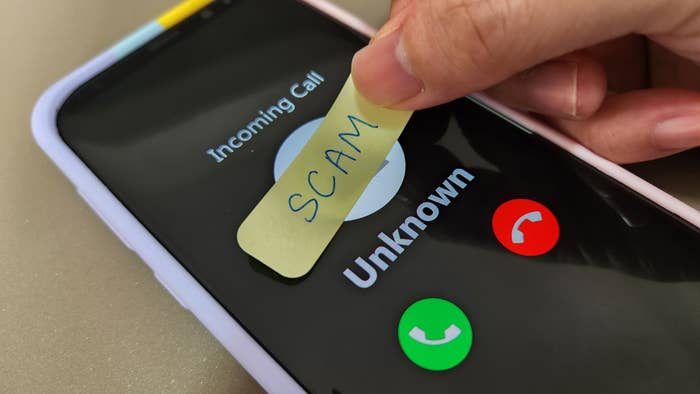Hand holding a sticky note labeled &quot;SCAM&quot; over a smartphone screen showing an incoming call from &quot;Unknown&quot; with options to accept or decline the call