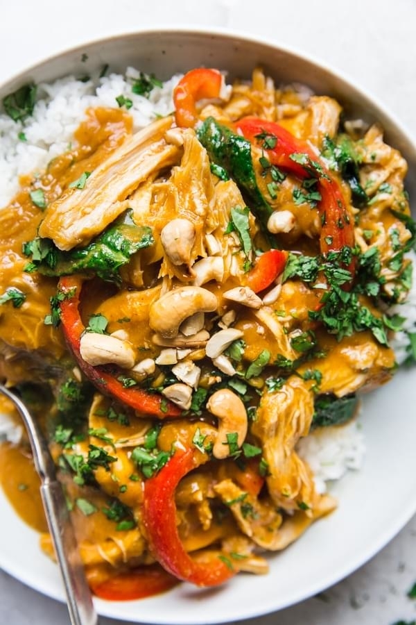A bowl of rice topped with Thai curry, featuring sliced chicken, red bell peppers, cashews, and chopped cilantro. A fork rests on the edge of the bowl