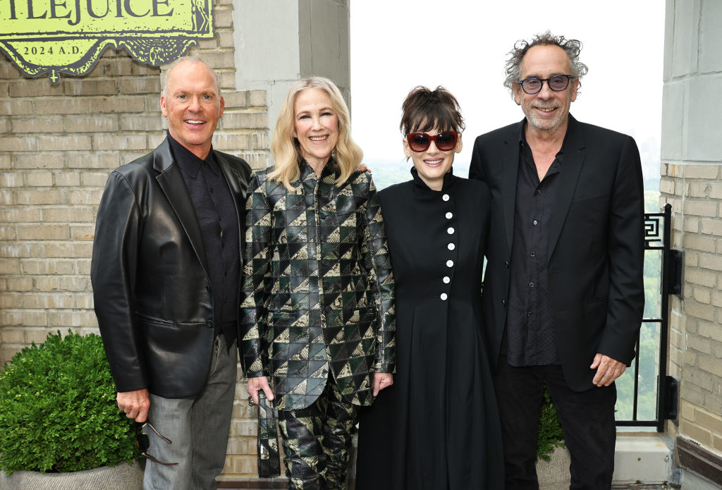 Michael Keaton, Catherine O'Hara, Winona Ryder, and Tim Burton pose together, Keaton in a leather jacket, O'Hara in a patterned suit, Ryder in dark dress, and Burton in a dark suit