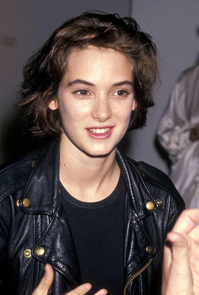 Winona Ryder smiles, wearing a dark leather jacket and a dark top