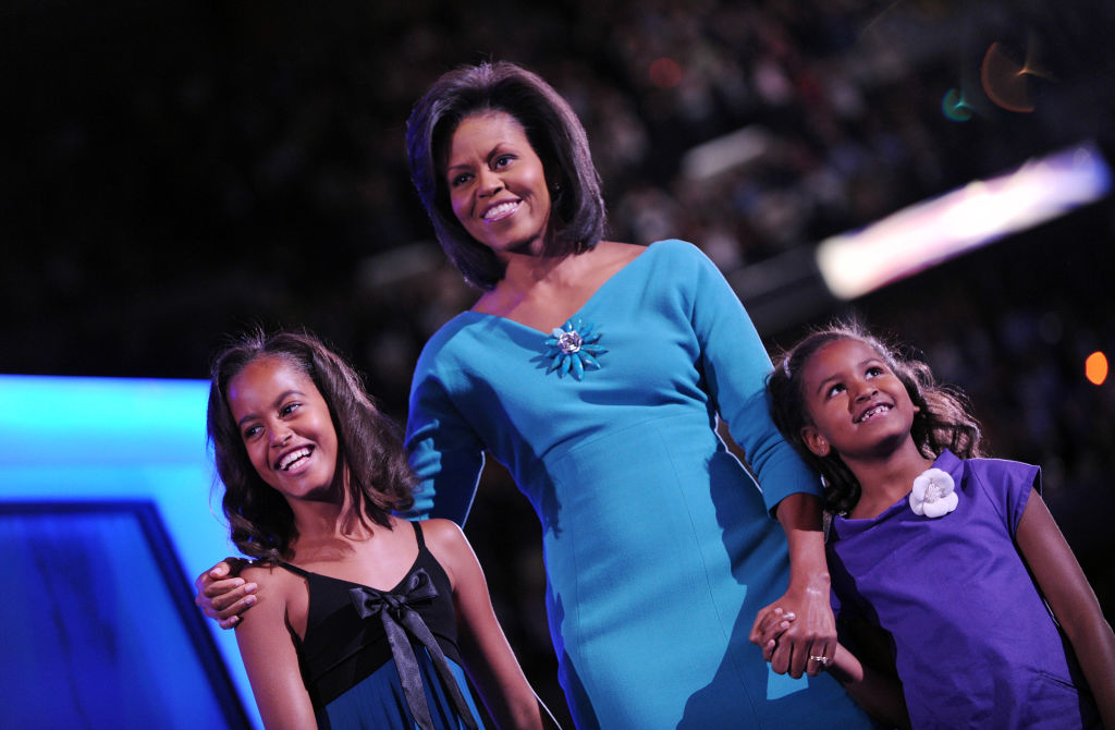 Michelle Obama stands smiling with her daughters, Sasha and Malia, dressed in elegant outfits at a public event