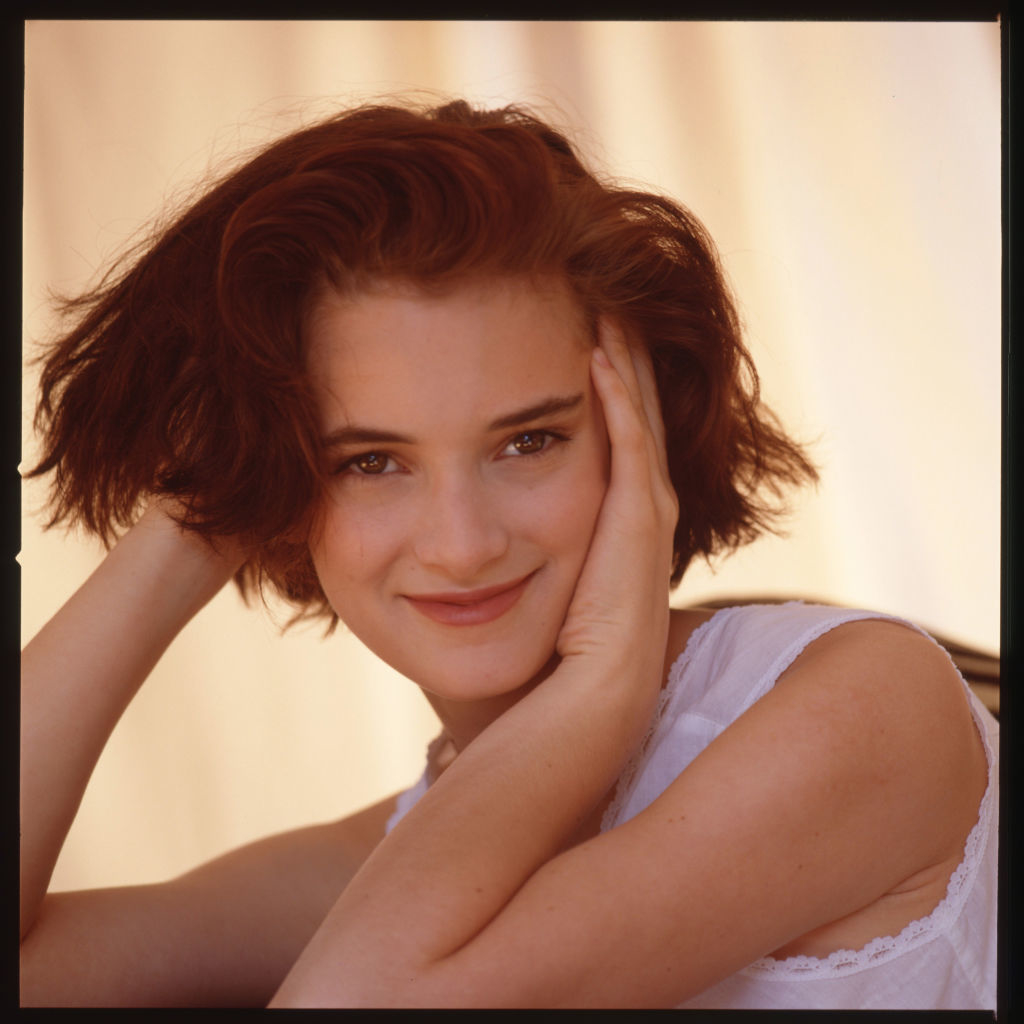 Winona Ryder smiles with hands on her cheeks, wearing a sleeveless white top against a light background