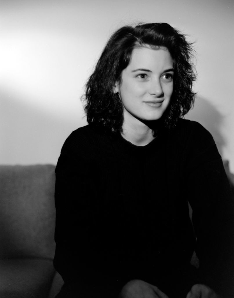 Winona Ryder in a black sweater, sitting on a couch, looking towards the camera