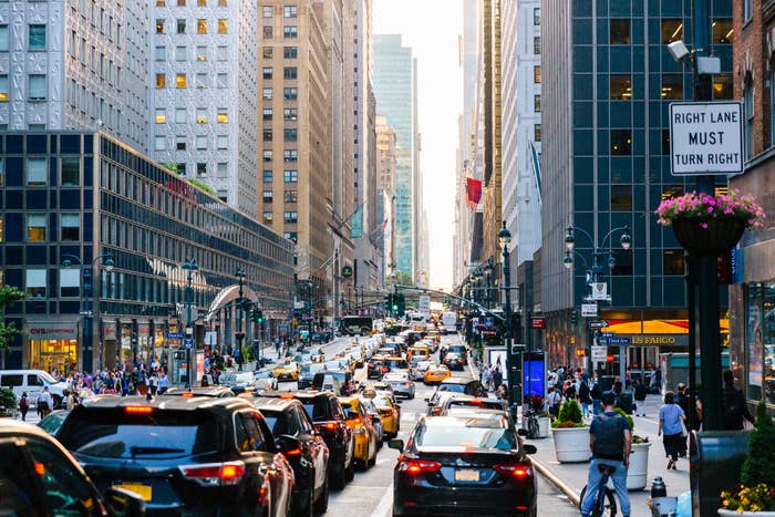 Busy city street with heavy traffic, pedestrians on sidewalks, and tall buildings on either side. Sign on right reads &quot;Right Lane Must Turn Right&quot;