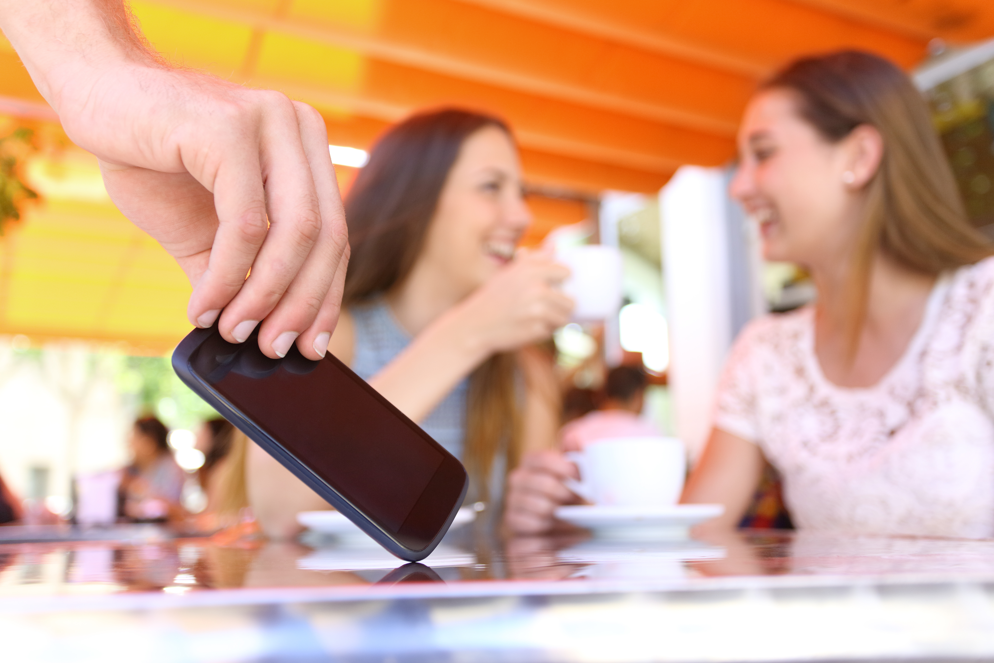 A hand reaches for a phone on a cafe table while two women in the background, out of focus, smile and converse over coffee