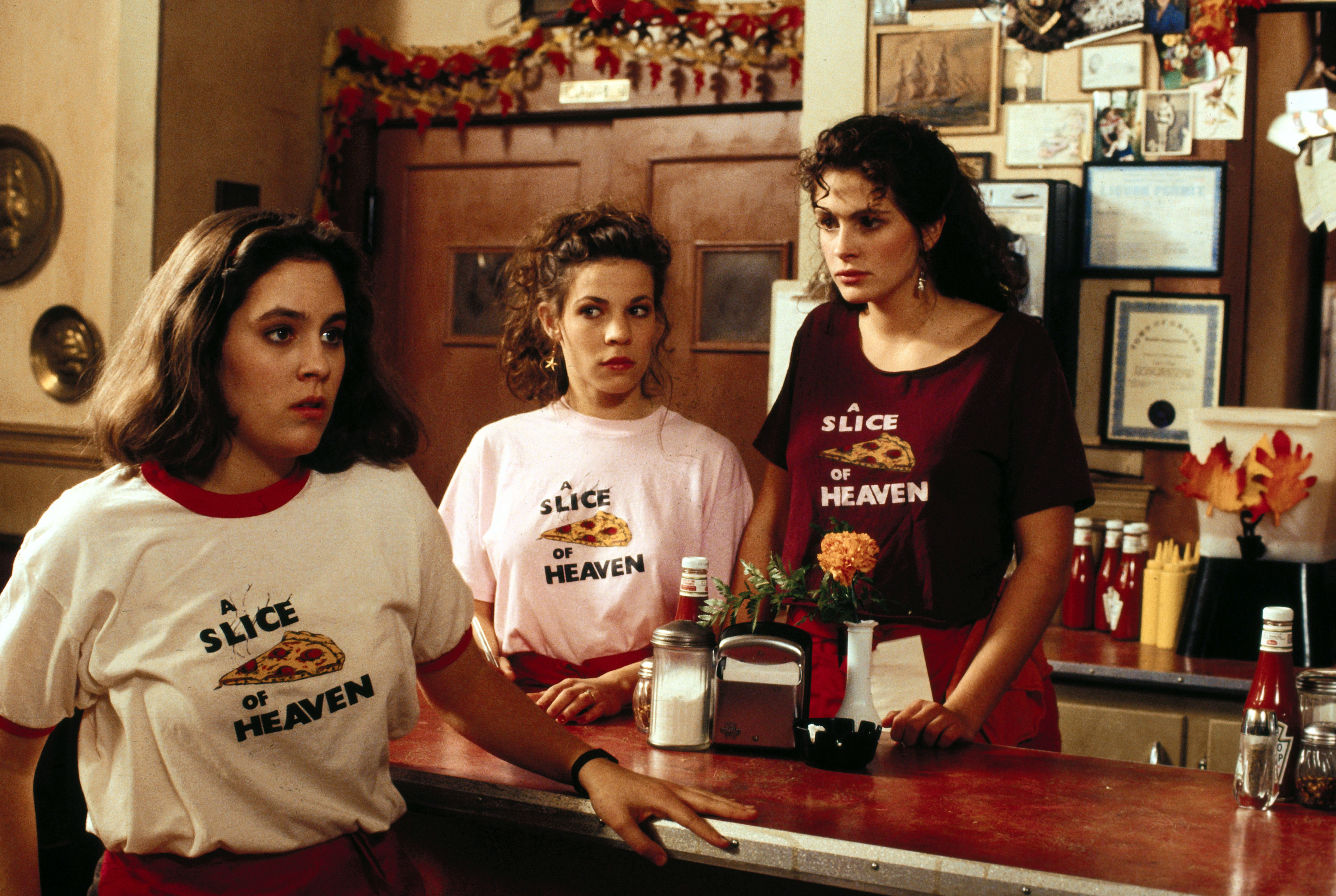 Three women, likely waitresses, are standing behind a diner counter wearing &quot;A Slice of Heaven&quot; t-shirts. The setting has a cozy, rustic aesthetic
