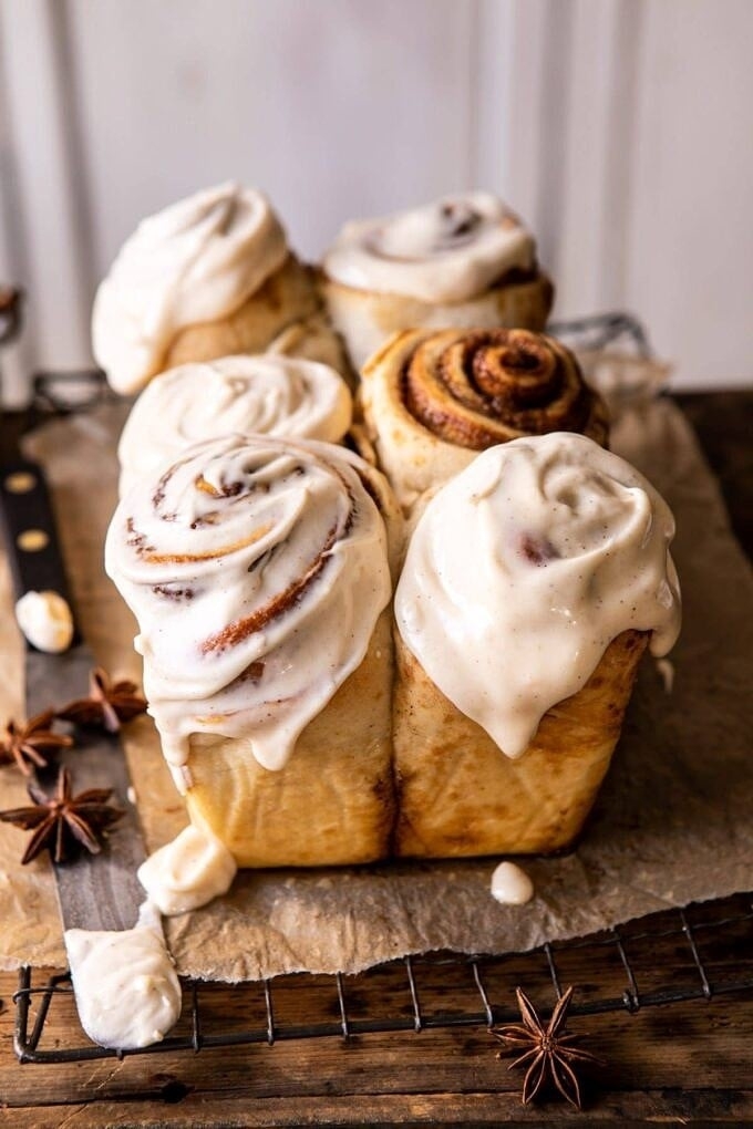 A tray of cinnamon rolls with thick icing, some with more icing and some slightly browned on top, displayed on parchment paper with star anise beside it