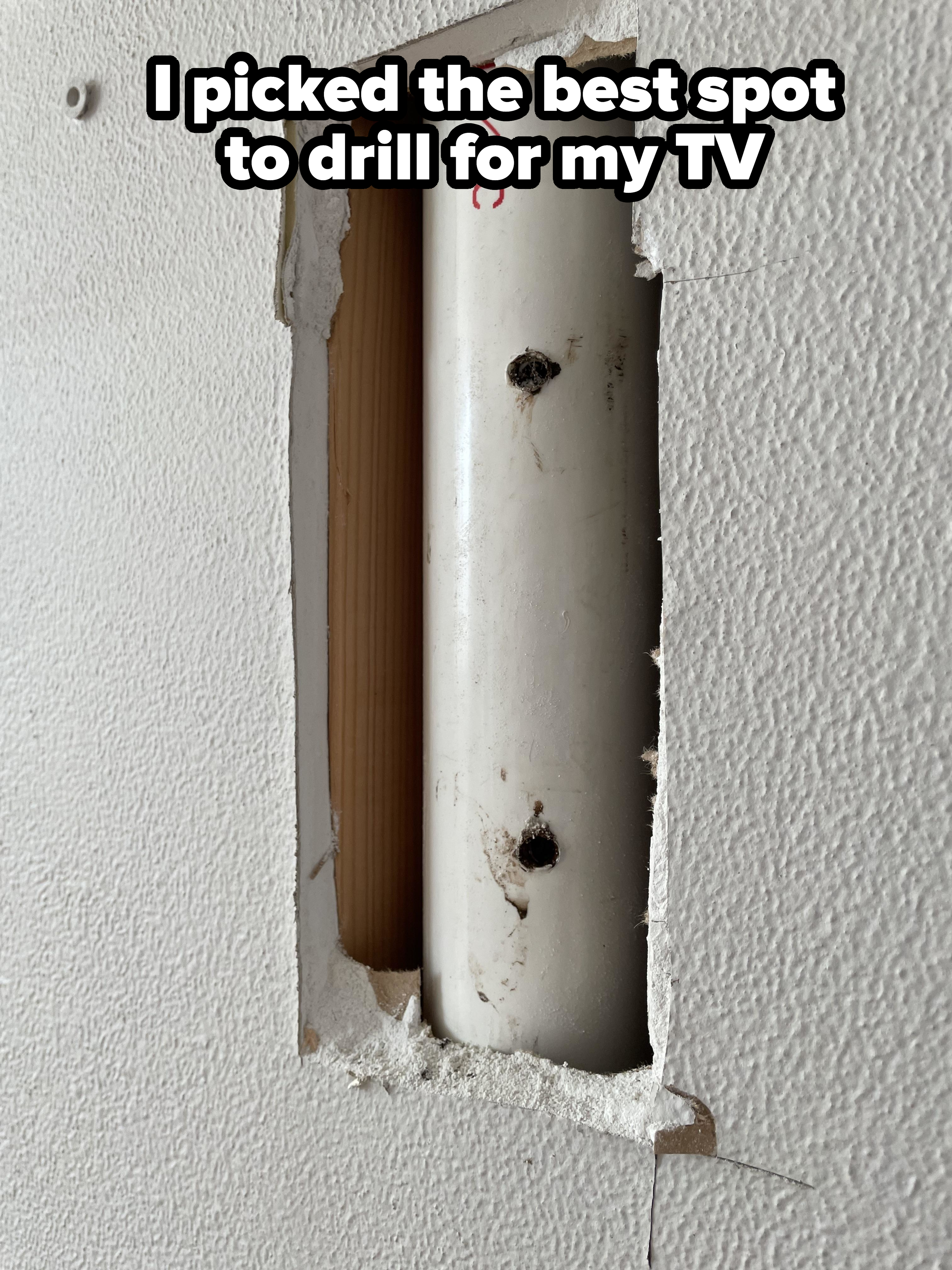 A wall with a rectangular hole exposing a white PVC pipe and a wooden stud. The pipe has red text and two screw holes