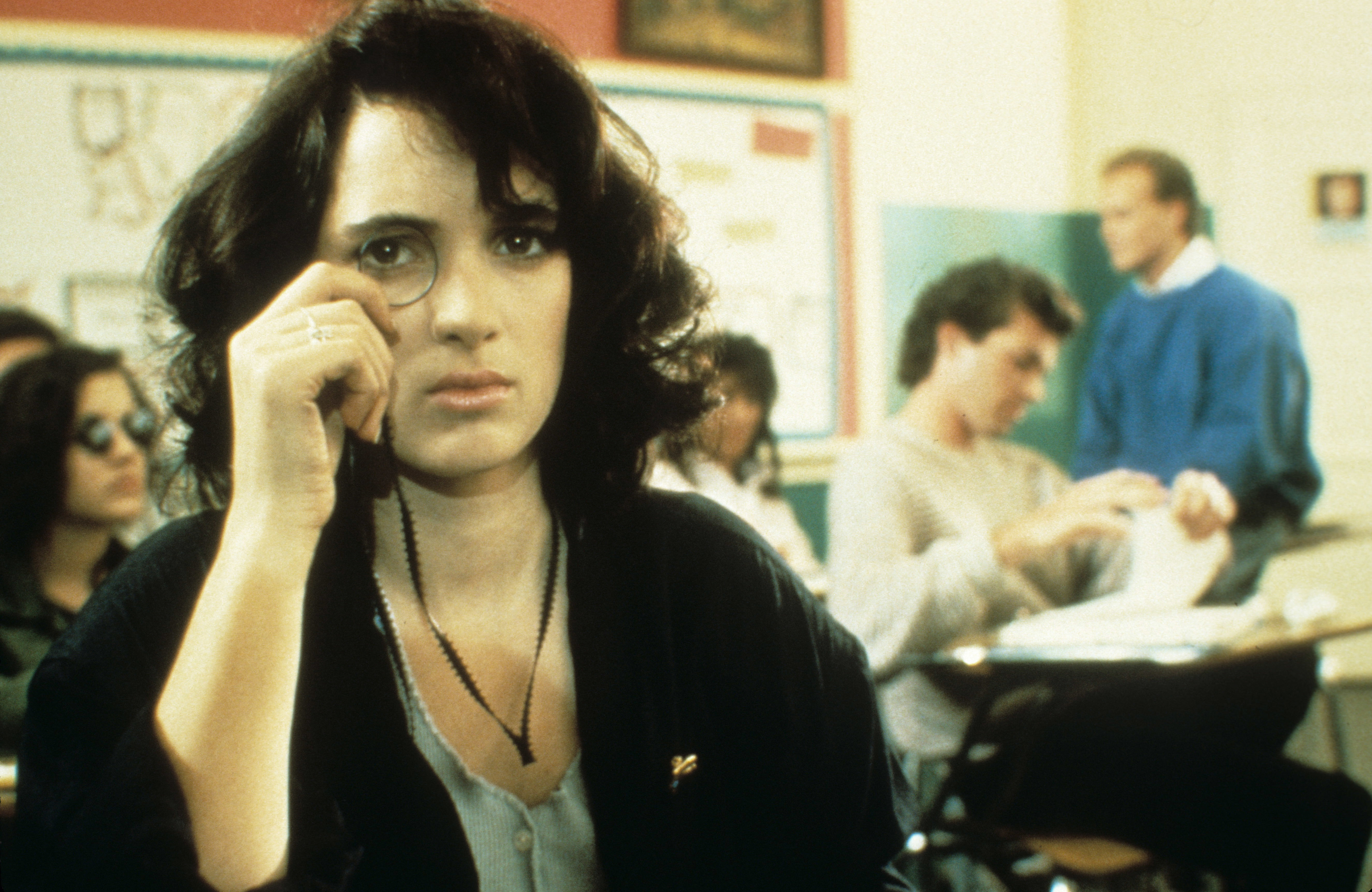 Winona Ryder as Veronica Sawyer in &quot;Heathers,&quot; dressed in school attire, holding a monocle, with classroom scenes in the background
