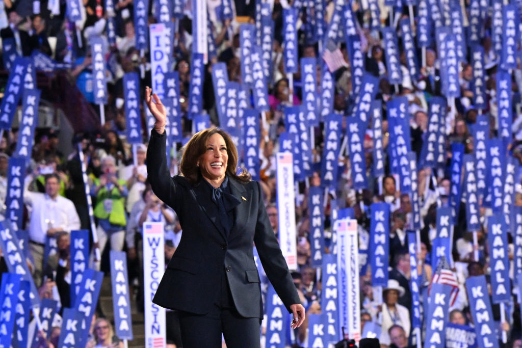 Kamala Harris waves while standing in front of a large crowd holding many &quot;KAMALA&quot; signs at a political event