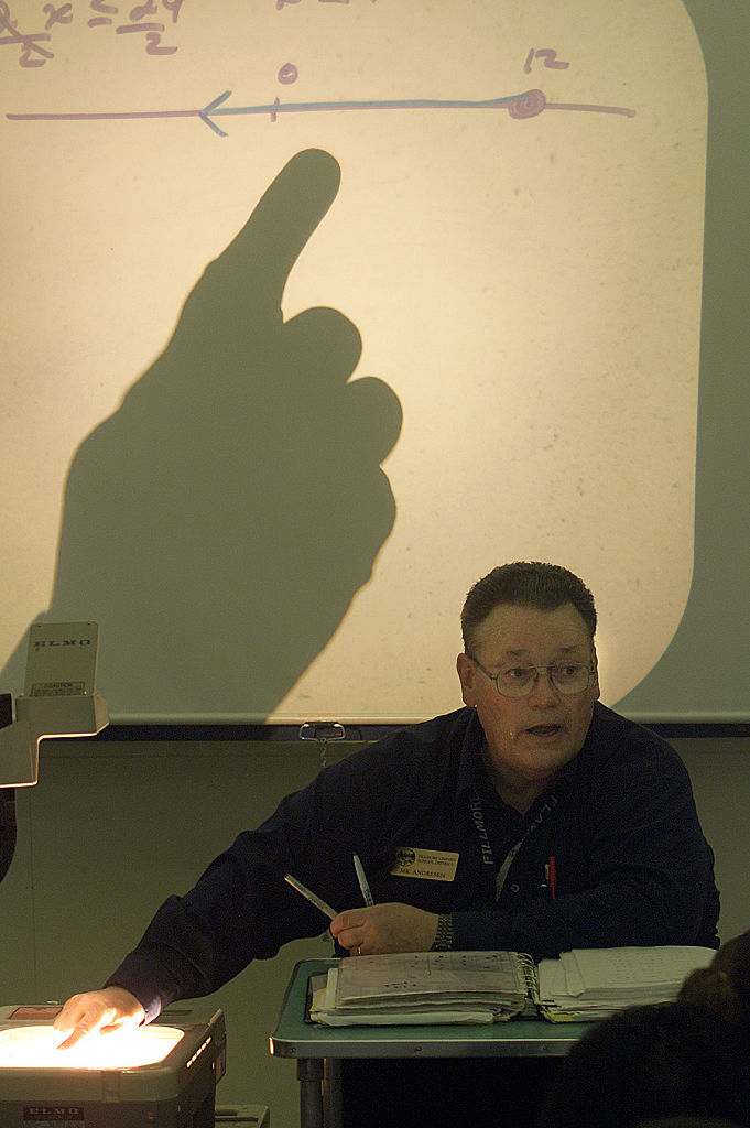 A person is seen writing on a paper with an overhead projector displaying a chart and shadows of hands. Several people are seated in the background