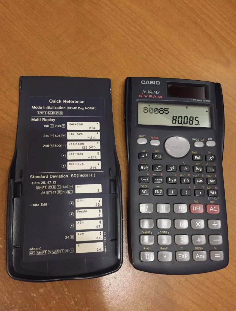 A Casio fx-300MS scientific calculator on a wooden surface, displaying &quot;80085&quot;. The calculator cover shows quick reference guides for various mathematical functions