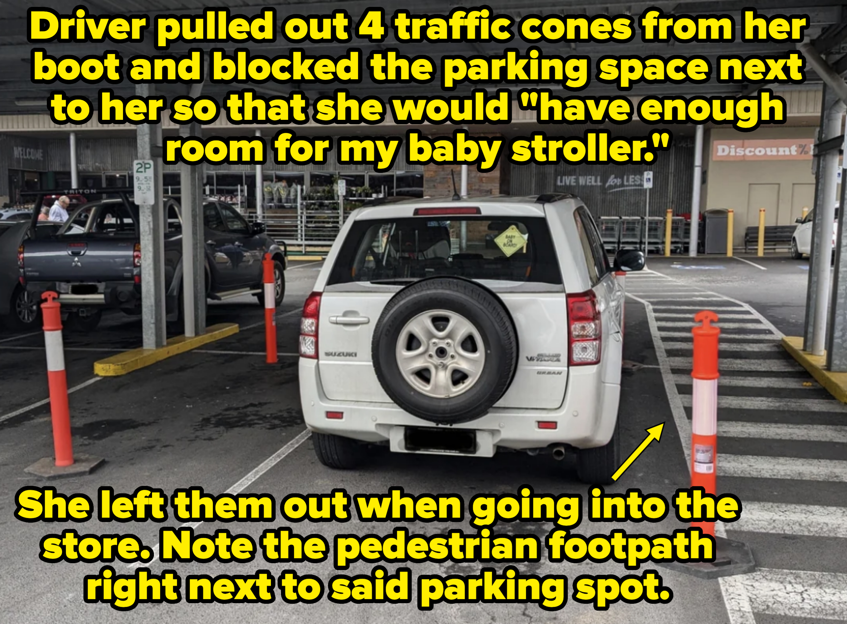 A white Suzuki SUV is parked in a covered parking spot with orange pylons around it in a partially filled lot outside a retail store