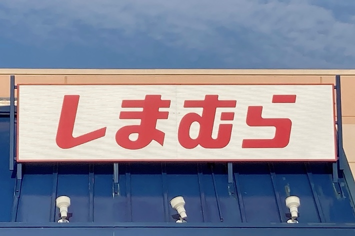 A retail store with a Japanese sign that reads &ldquo;しまむら&rdquo; above the entrance. There is a handicapped parking sign visible in the foreground