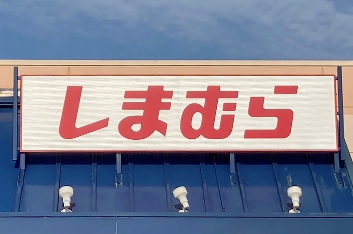 The image shows the exterior of a store with a sign in Japanese that reads &ldquo;しまむら&rdquo; above the entrance. A handicap parking sign is visible