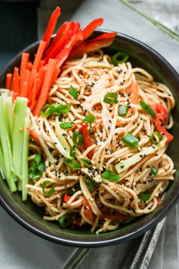 Bowl of noodles topped with sesame seeds, sliced green onions, and garnished with carrot and cucumber sticks