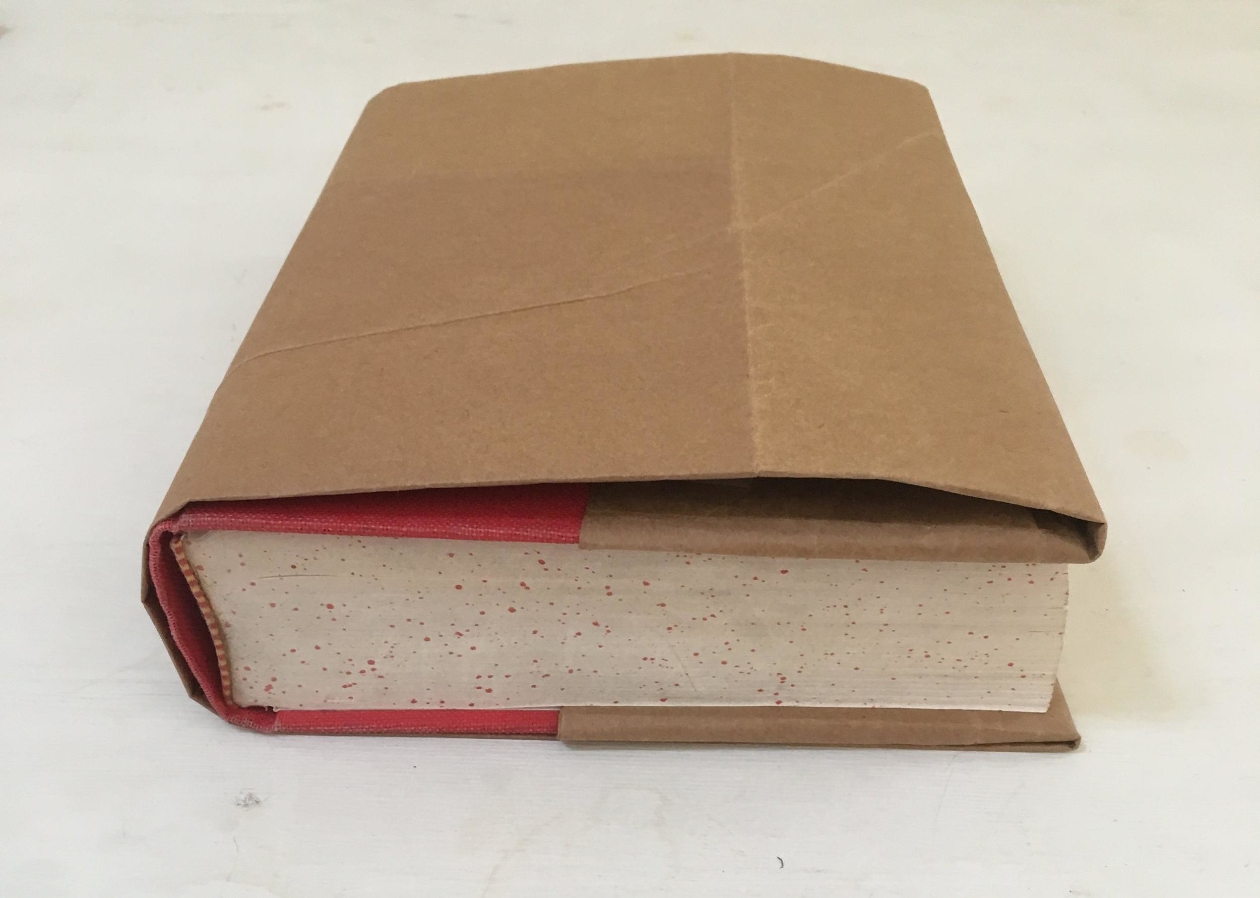 A thick book with red edges is wrapped in a plain brown paper cover, revealing only the spine and edge of the pages