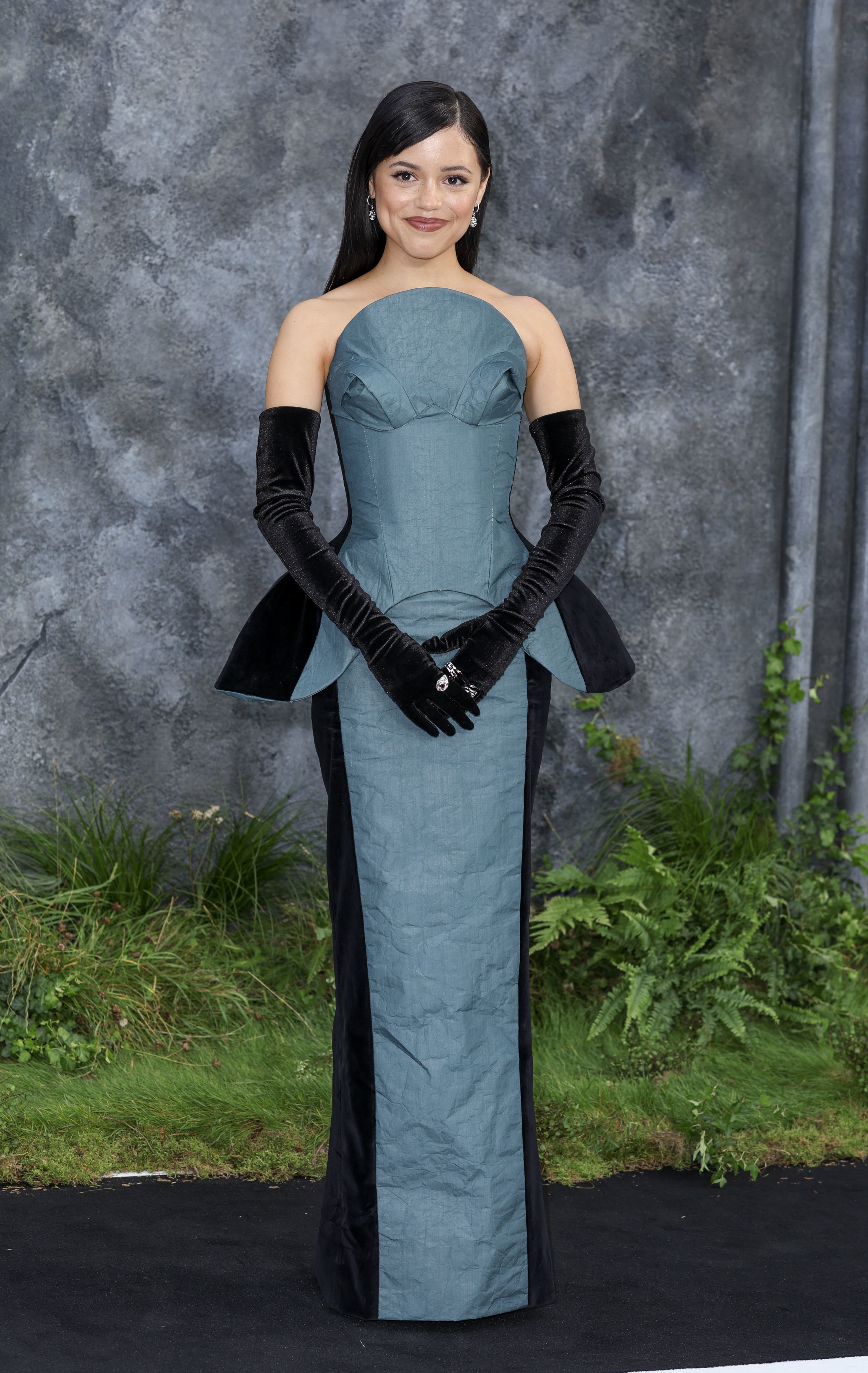 Jenna Ortega stands in an elegant, strapless, structured gown with dramatic opera gloves at a formal event. Greenery and a textured wall are in the background