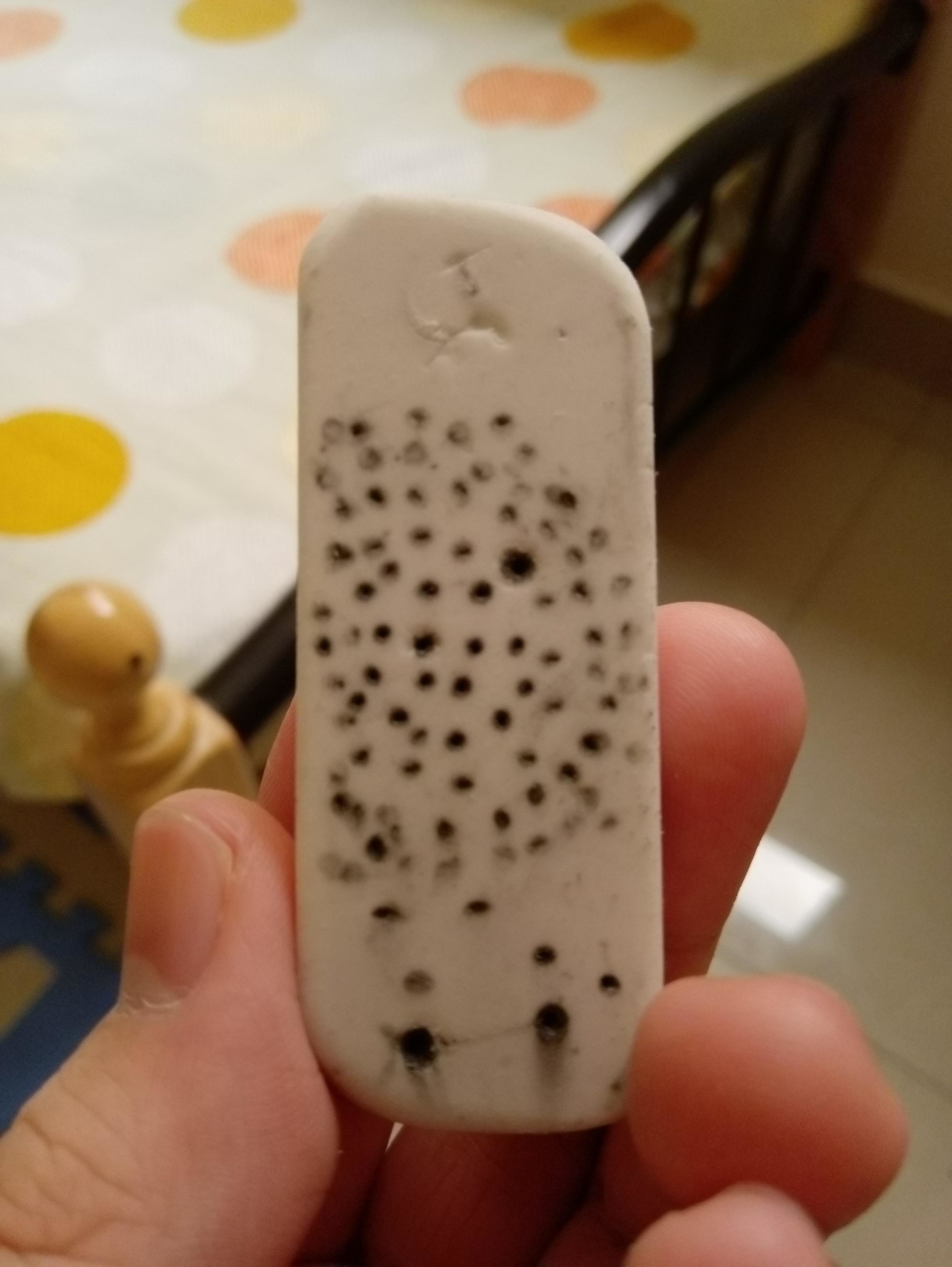 Hand holding a well-used eraser with numerous small black dots and markings, with the background showing part of a bed