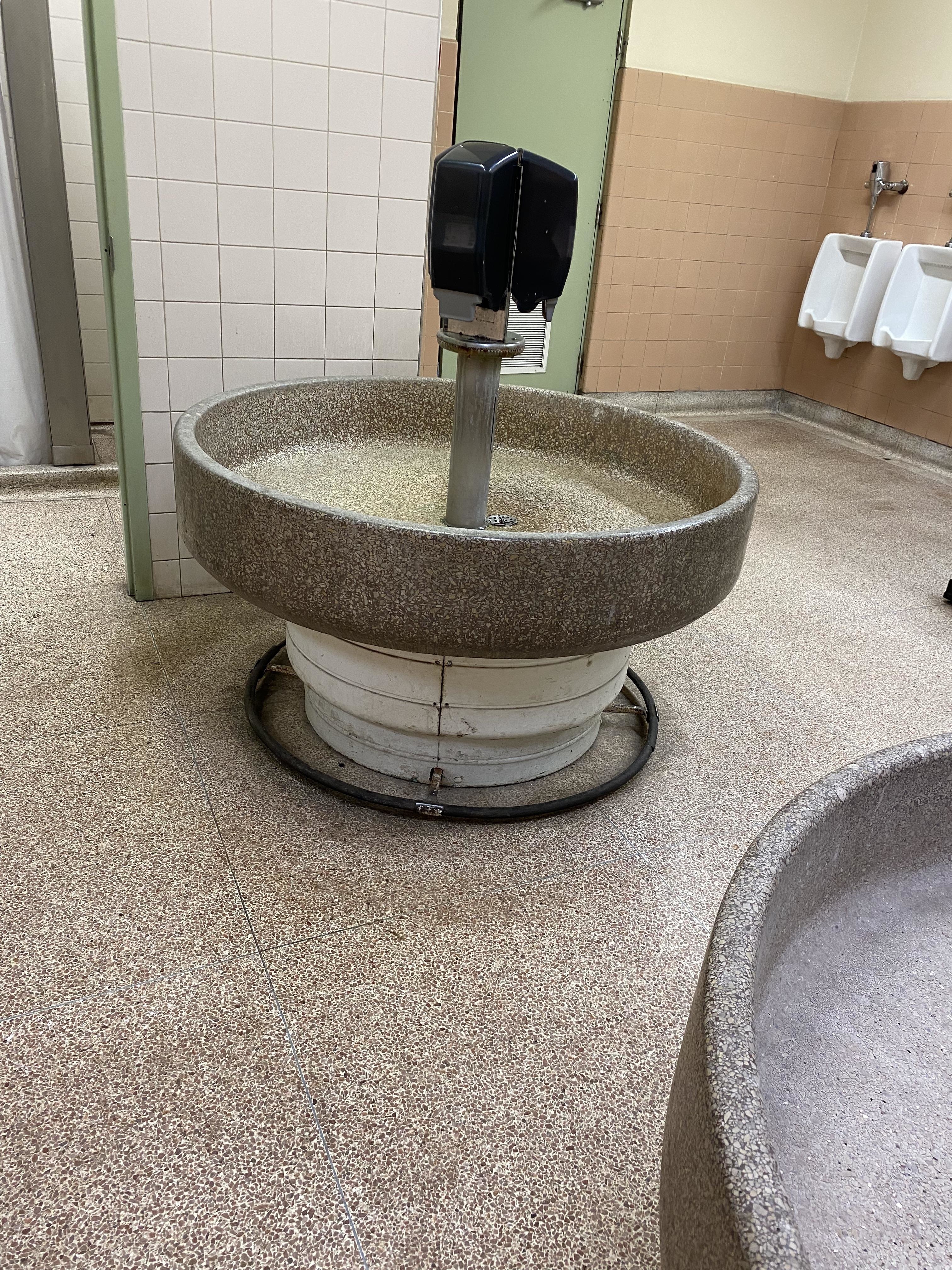 A round, stone-wash bathroom sink with a hands-free soap dispenser in a public restroom. Urinals and tiled walls are visible in the background