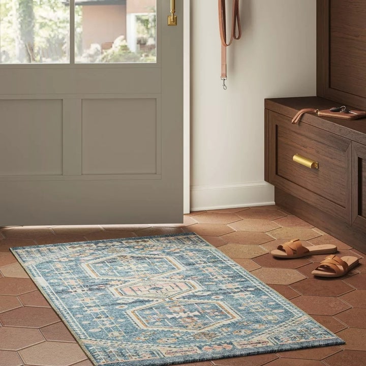 Patterned rug on hexagonal tiled floor near an entryway with a door, hooks for keys, and a shoe bench with leather sandals beside it