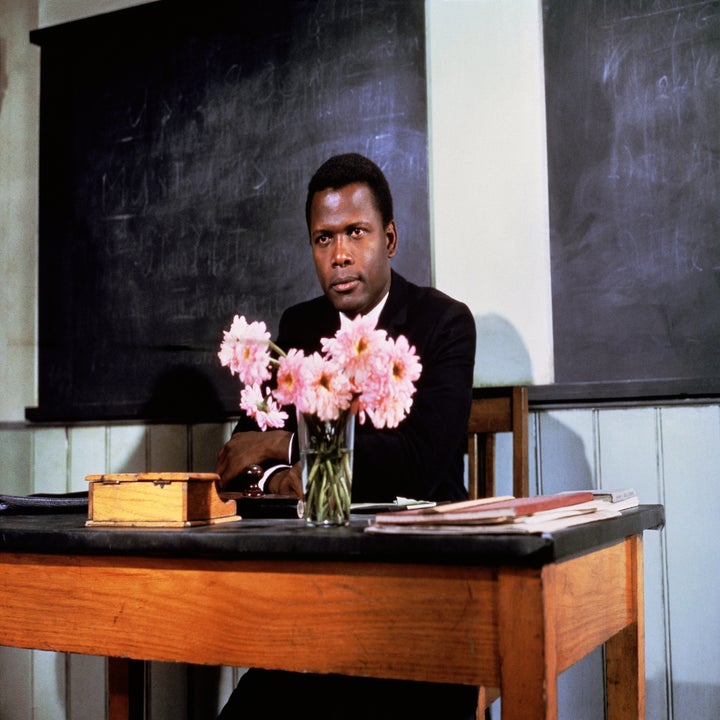 Sidney Poitier in a classroom setting, sitting at a desk with a vase of pink flowers, books, and a box, with chalkboards in the background