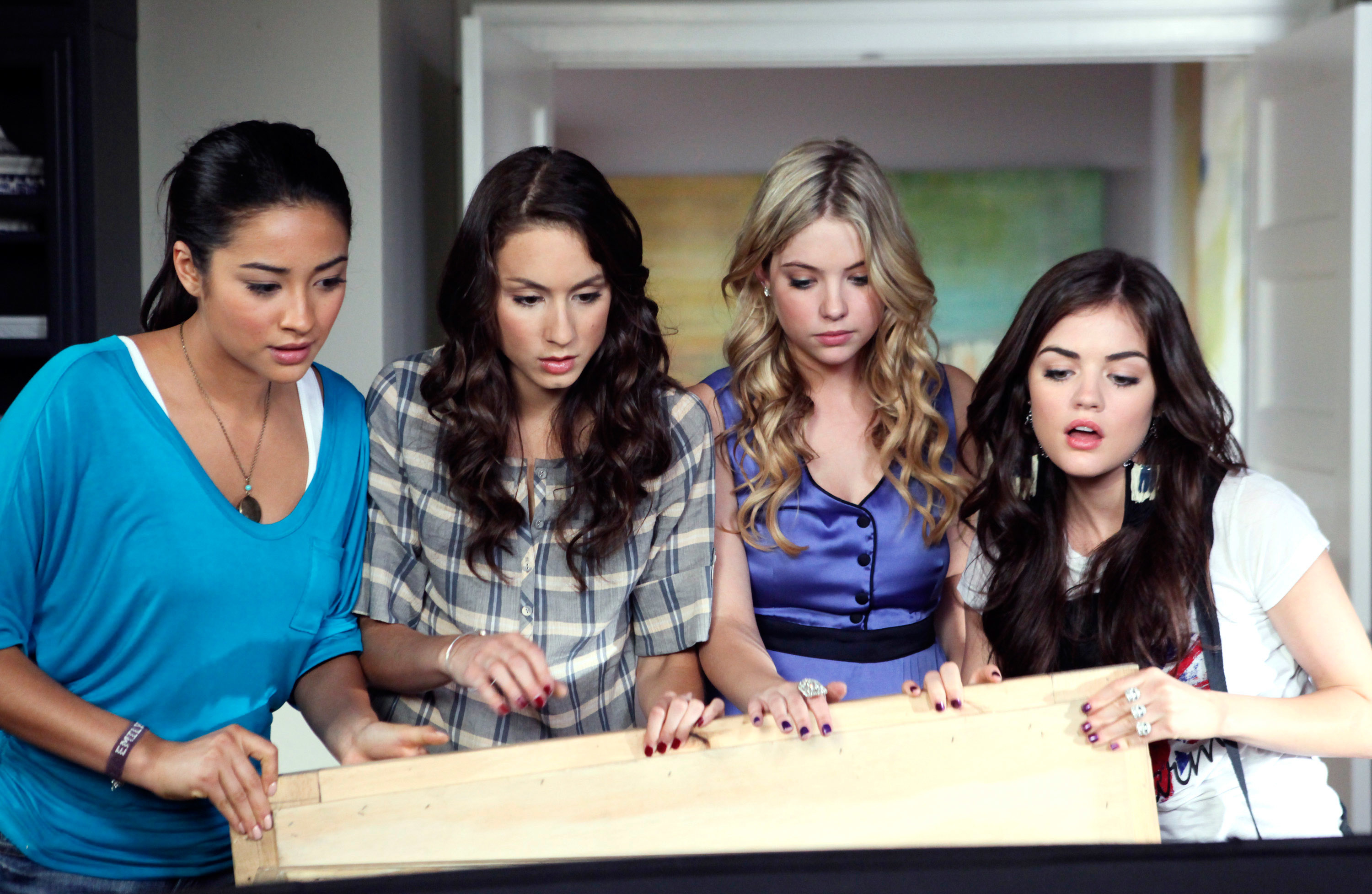 Shay Mitchell, Troian Bellisario, Ashley Benson, and Lucy Hale are gathered, looking at an object off-camera with serious expressions in a scene from a TV show