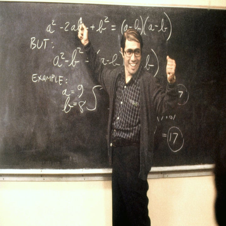 Jaime Escalante, wearing a cardigan over a plaid shirt, stands in front of a blackboard with algebraic equations, smiling and giving two thumbs up