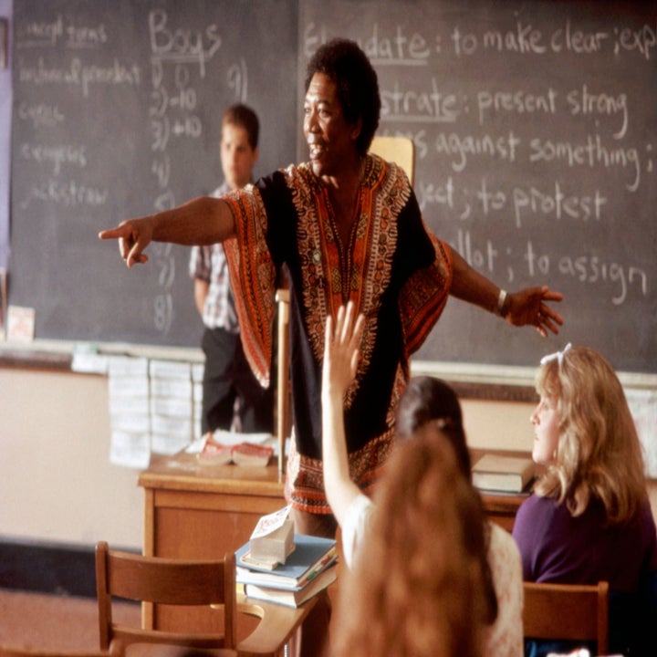 A teacher points to a raised hand of a student in a classroom. A chalkboard with educational notes is visible in the background