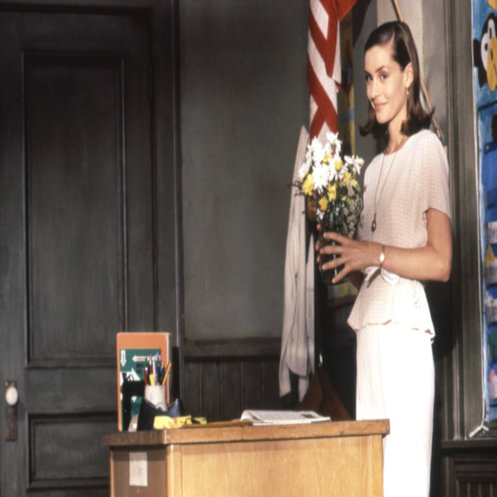 A woman in a light-colored outfit stands in a classroom holding a vase of flowers on a teacher's desk, with an American flag and bulletin board behind her