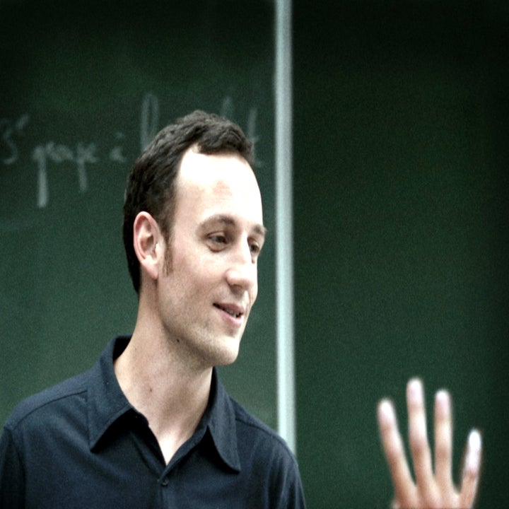 A man wearing a collared shirt stands in front of a chalkboard, engaged in conversation with another person whose hand is visible in the foreground