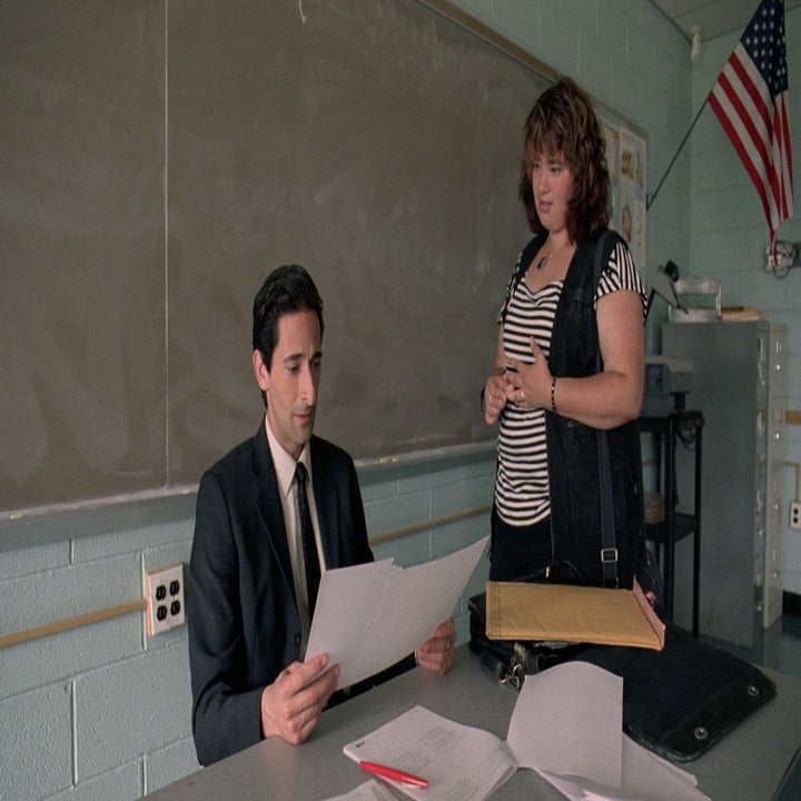 A man in a suit sits at a desk looking at papers while a woman in a striped shirt and black vest stands by, classroom setting, an American flag in the background