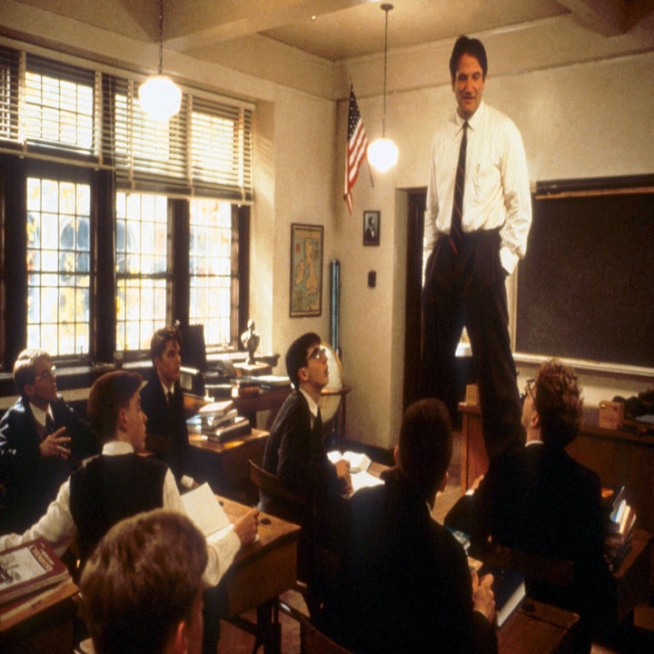 Robin Williams stands on a desk teaching students in a classroom, conveying an engaging and unconventional teaching style