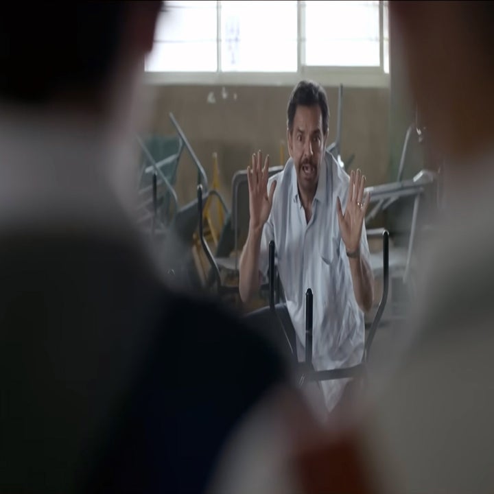 Scene from a movie where a man in a white shirt interacts animatedly with others, hands raised. The setting appears to be a room with scattered furniture.