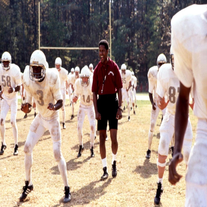 A football coach leads a team practice on a field. Players wear white uniforms with helmets