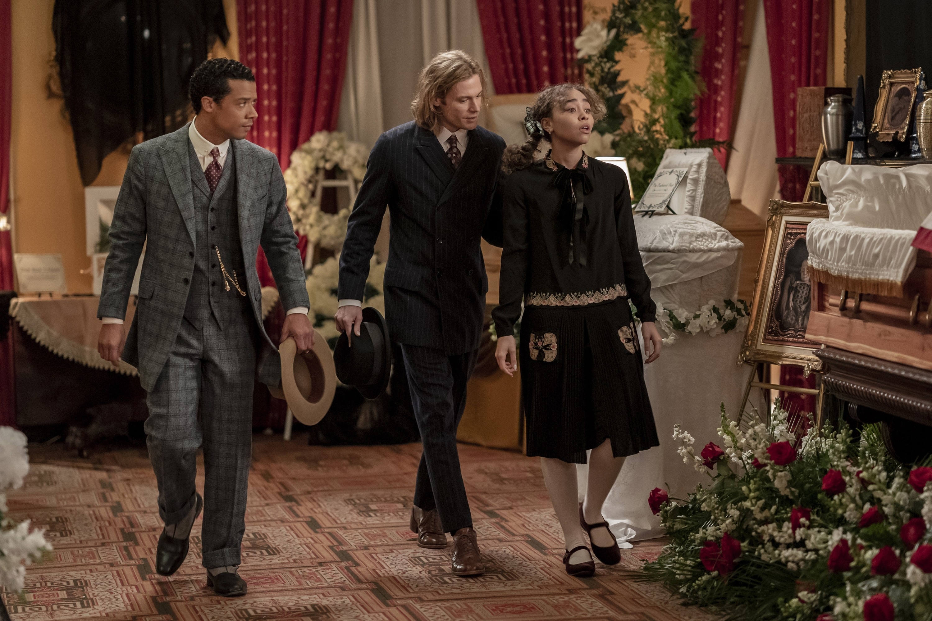 Jacob Anderson, Sam Reid, and Bailey Bass are in a period setting; Anderson in a suit, Reid holding a hat, and Bass in a dress with lace and floral accents