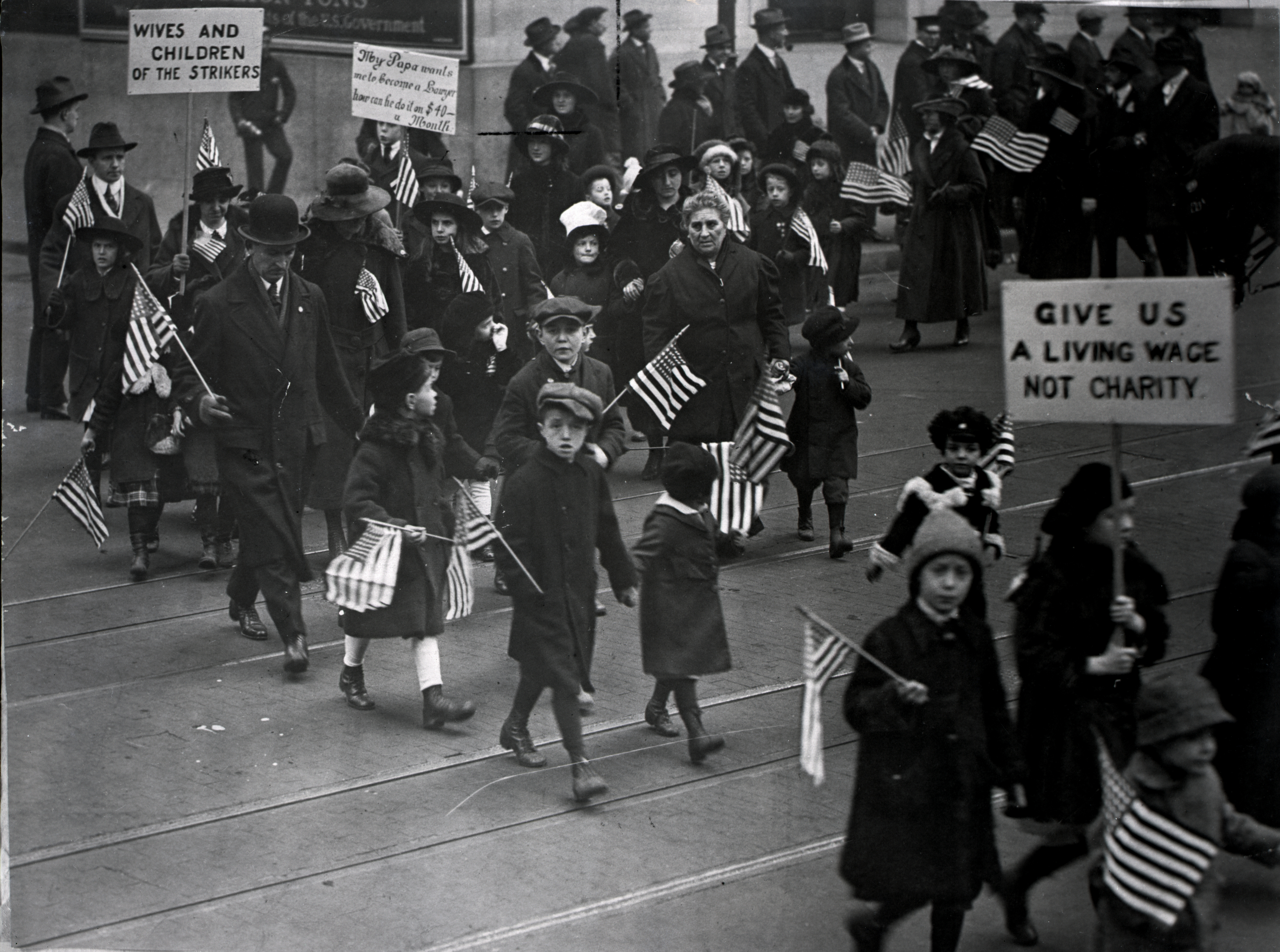 A vintage photo of a protest march with men, women, and children carrying American flags and signs, including "Wives and Children of the Strikers" and "Give Us a Living Wage Not Charity."