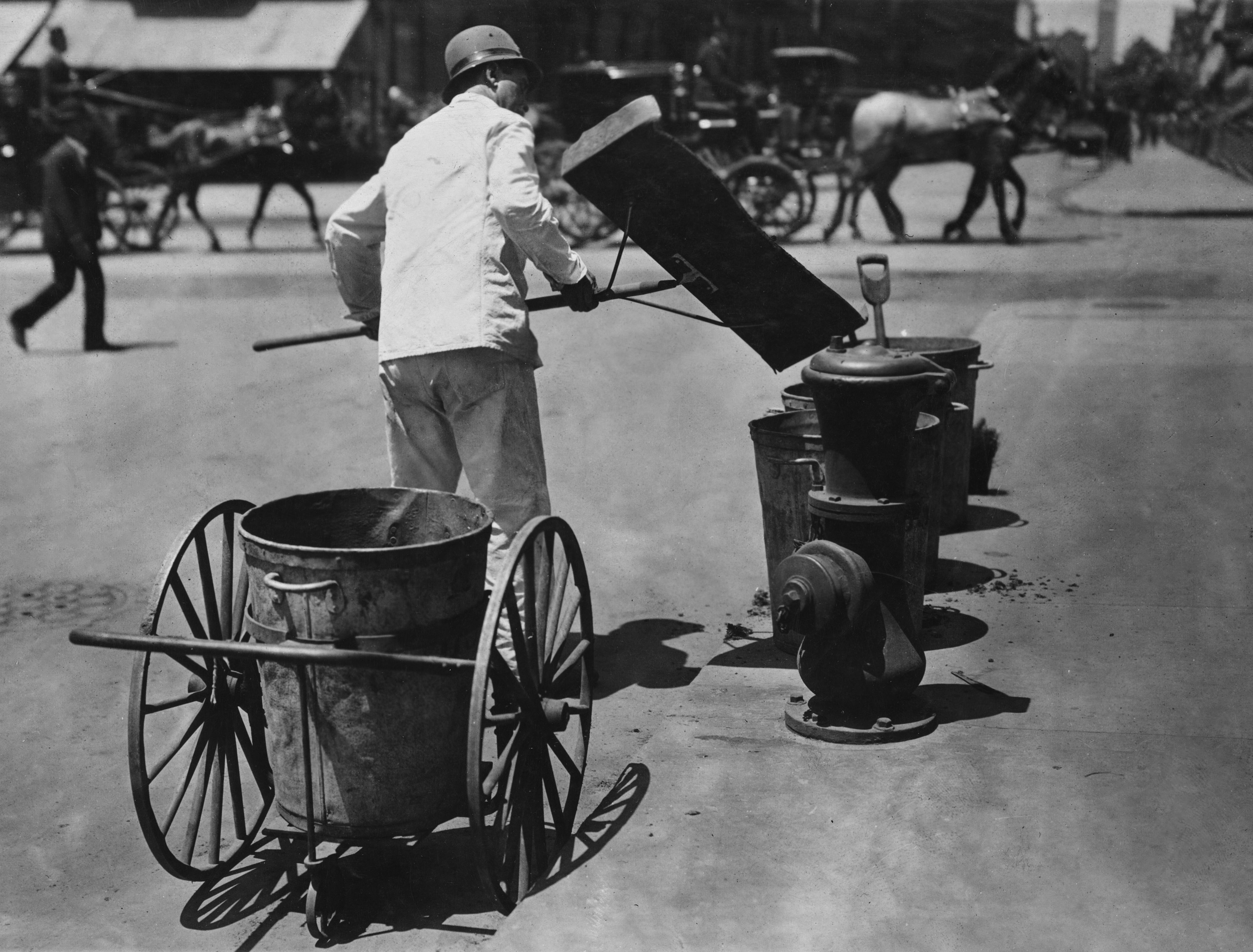 A person in work attire is emptying a street bin into a wheeled cart in an early 20th-century urban setting, with horse-drawn carriages in the background