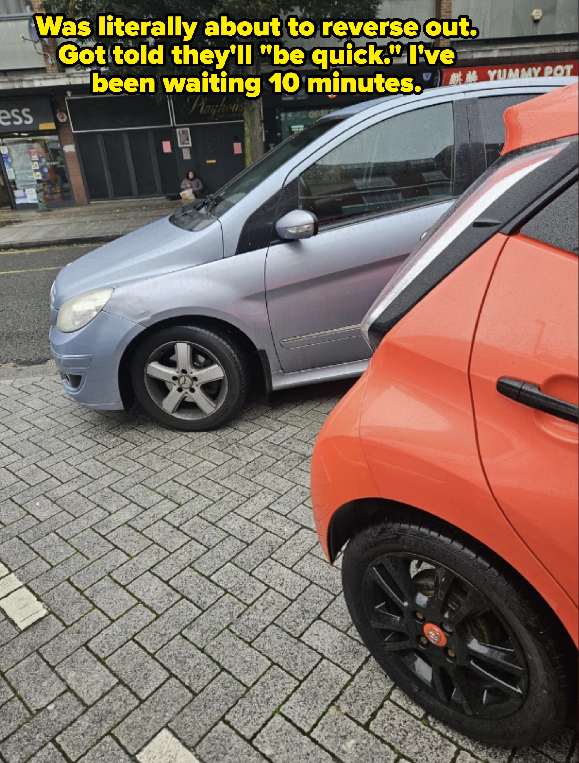 Two parked cars on a street; a blue car in front and an orange car behind. Visible storefronts and restaurants in the background