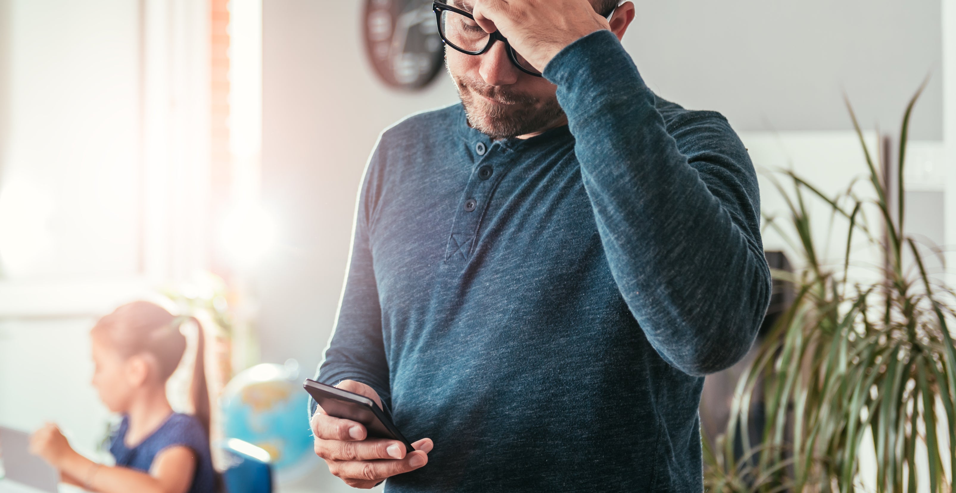 A man with glasses holds his forehead while looking at a phone. A child is at a desk in the background