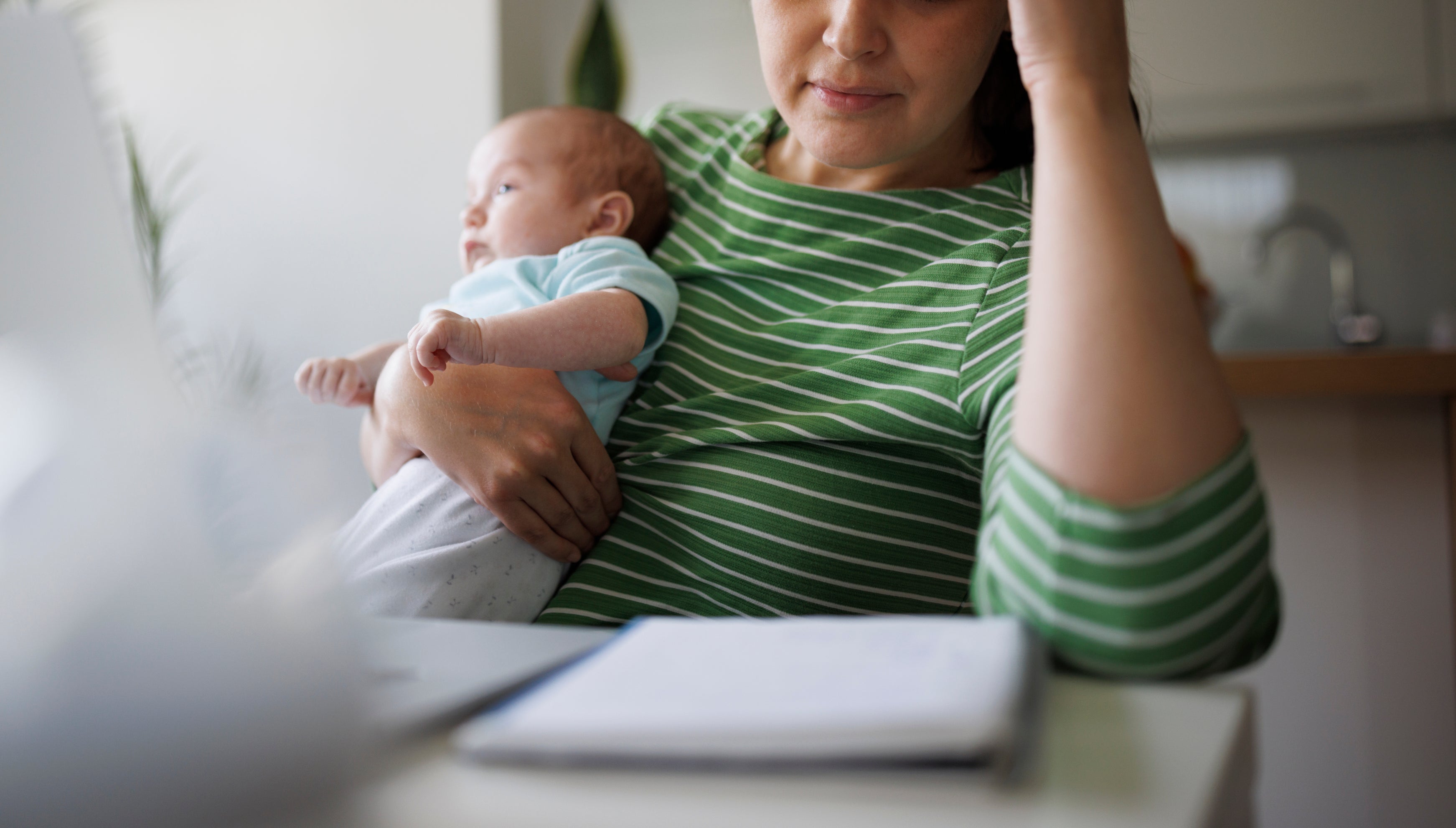 A woman holds a baby in one arm while working on a laptop and holding her head with the other hand, appearing stressed or tired