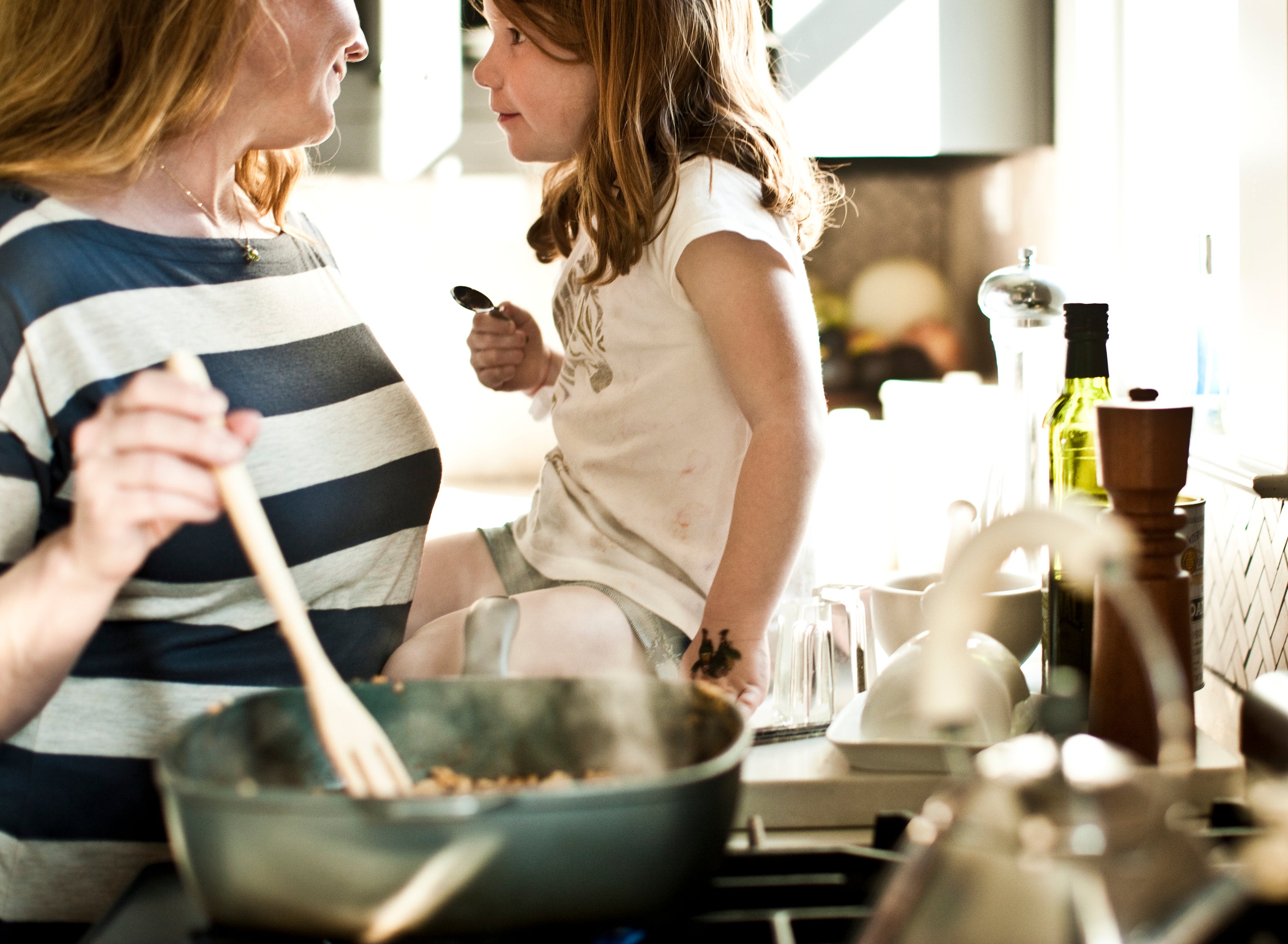 A woman and a young girl share a moment in the kitchen. The woman wears a striped shirt and the girl sits on the counter holding a spoon