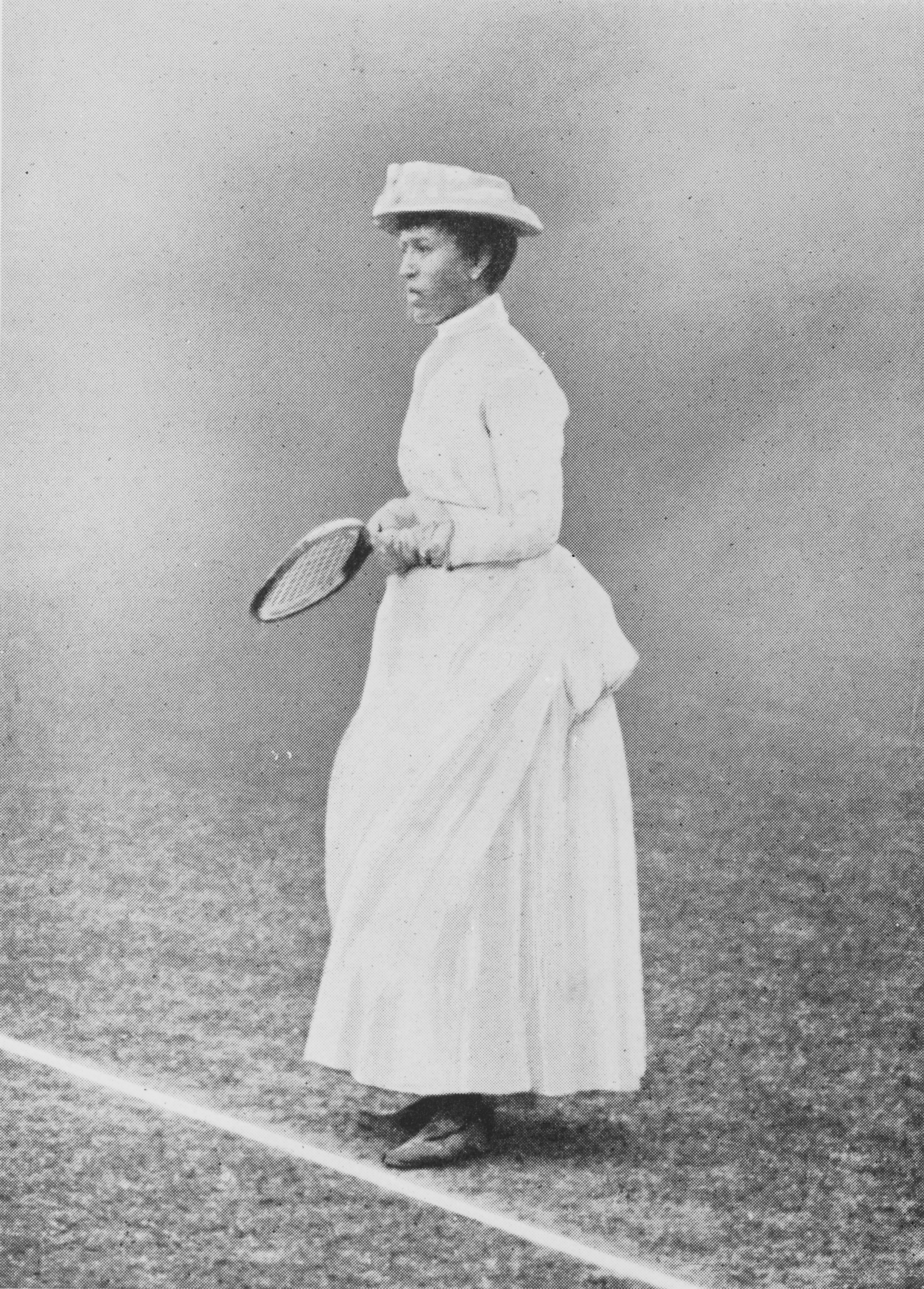 A woman stands on a tennis court in Victorian-style attire, including a hat and a long white dress, holding a tennis racket