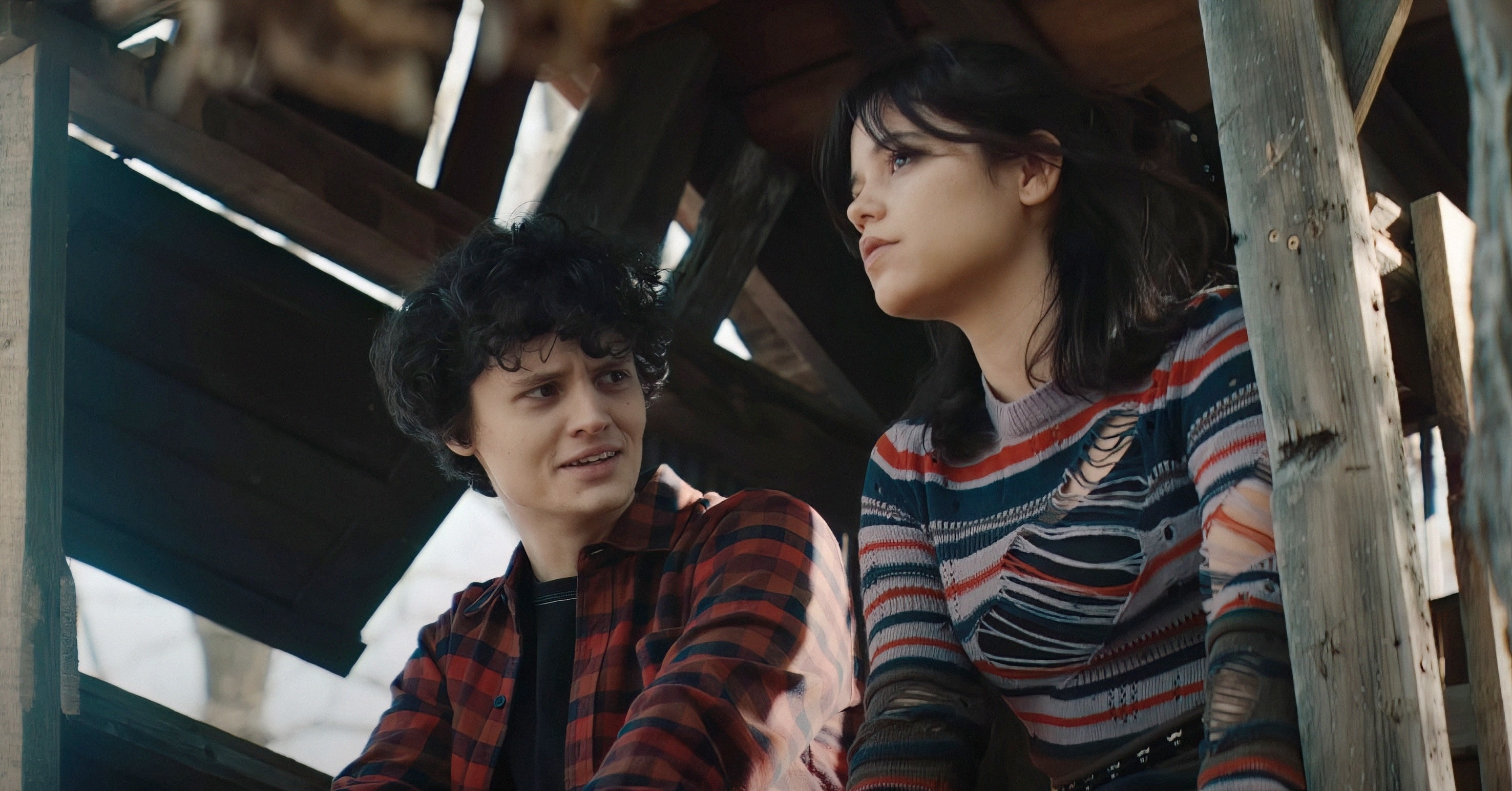 Percy Hynes White in a checkered shirt and Jenna Ortega in a striped top sit together in a wooden structure