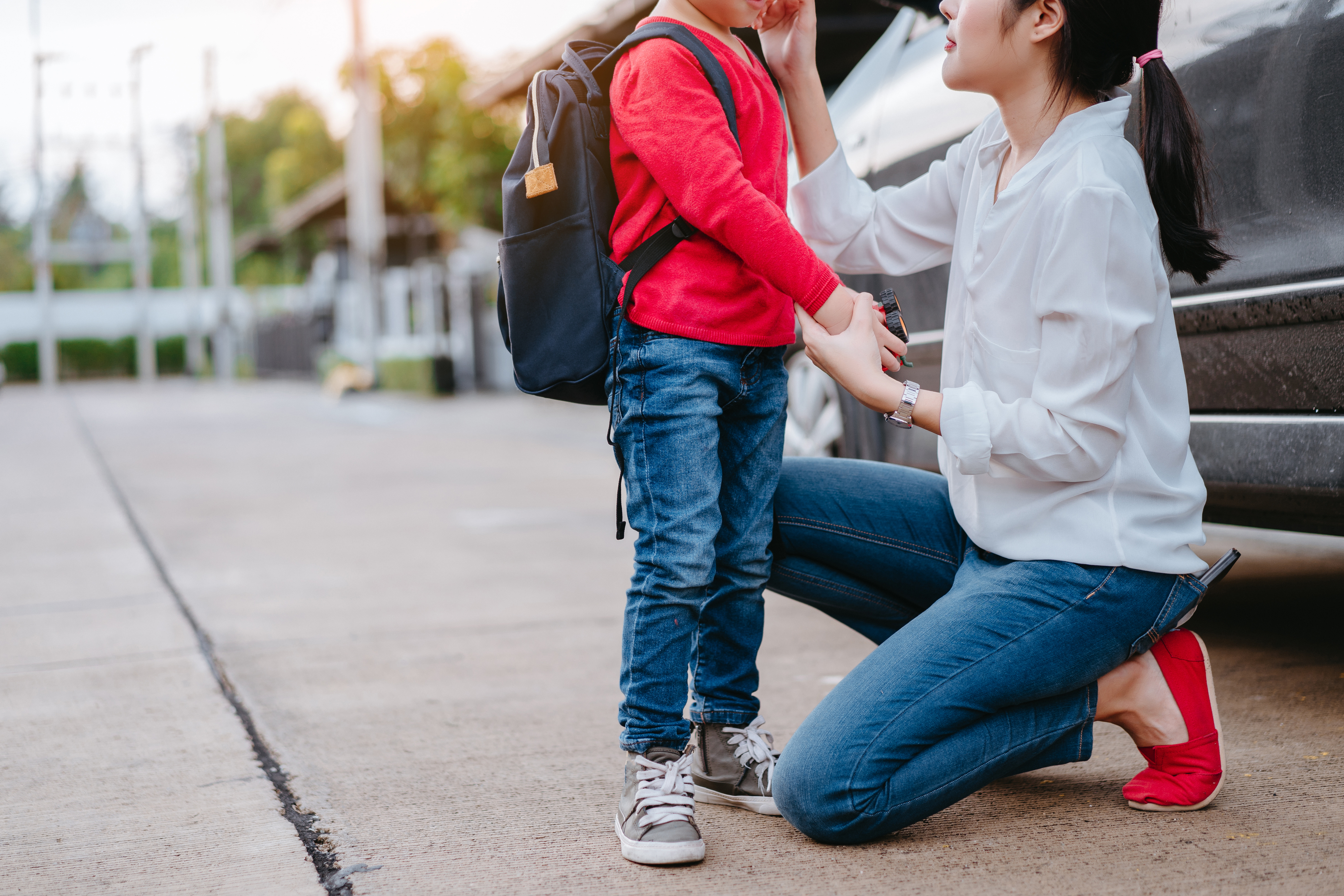 An adult kneels and adjusts a child's backpack. Both are casually dressed; the child wears jeans and sneakers