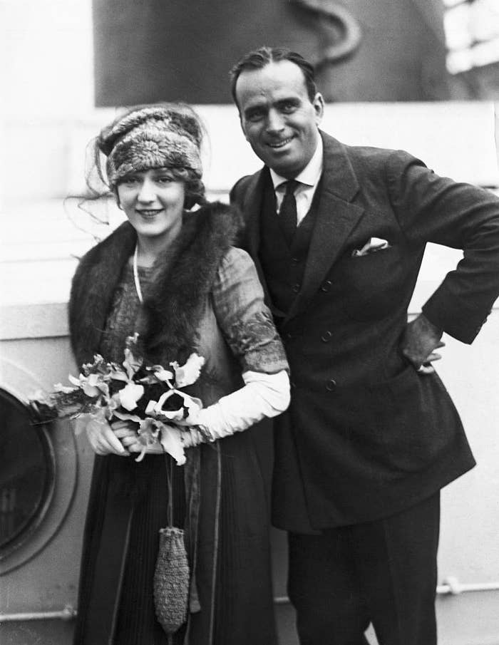 Two people, Mary Pickford in a fur stole and hat holding flowers, and Douglas Fairbanks in a suit, smiling and posing together