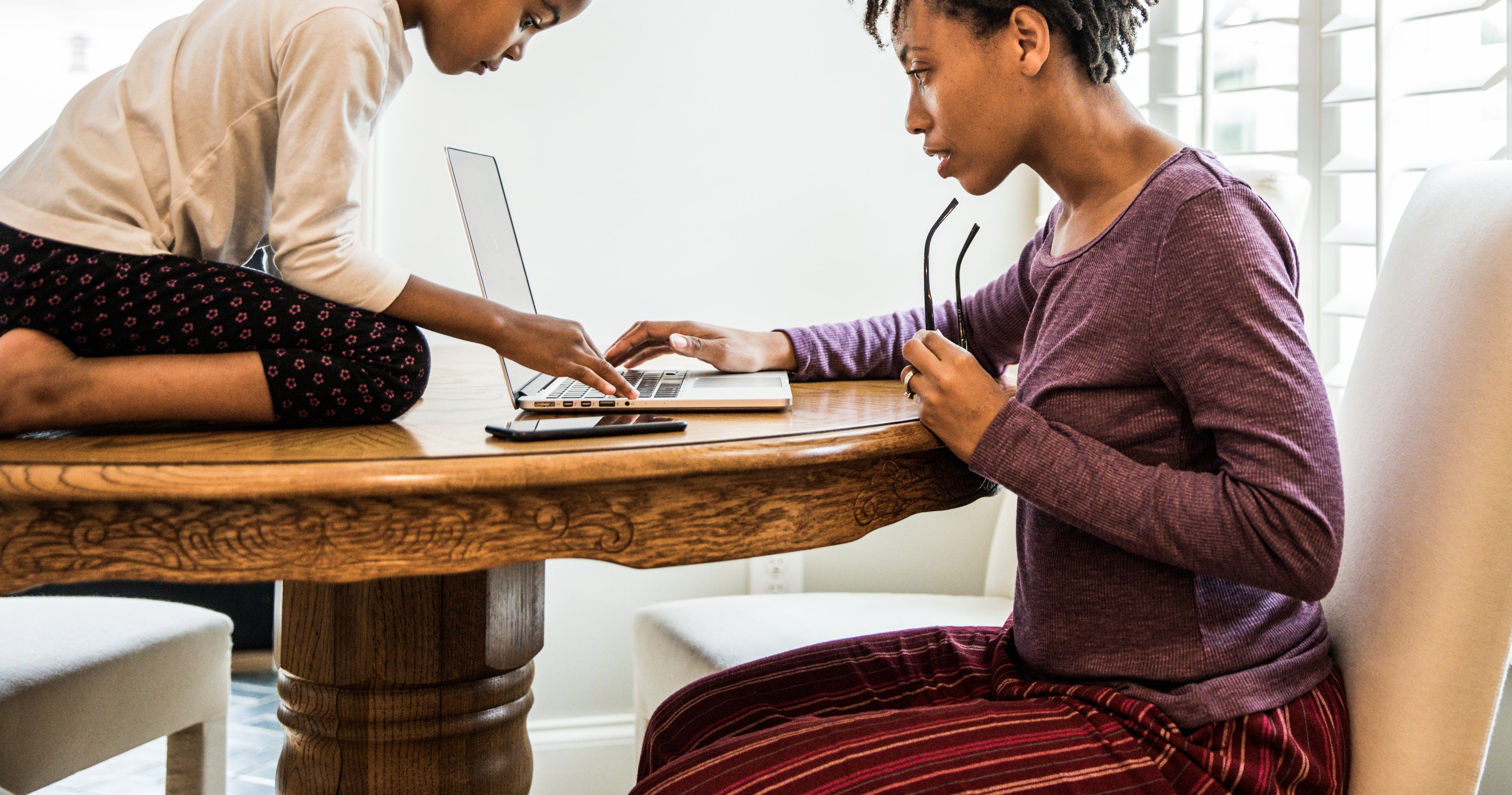 A young girl and an adult woman collaborate at a wooden table. The girl is focused on a laptop while the woman, holding glasses, is writing in a notebook