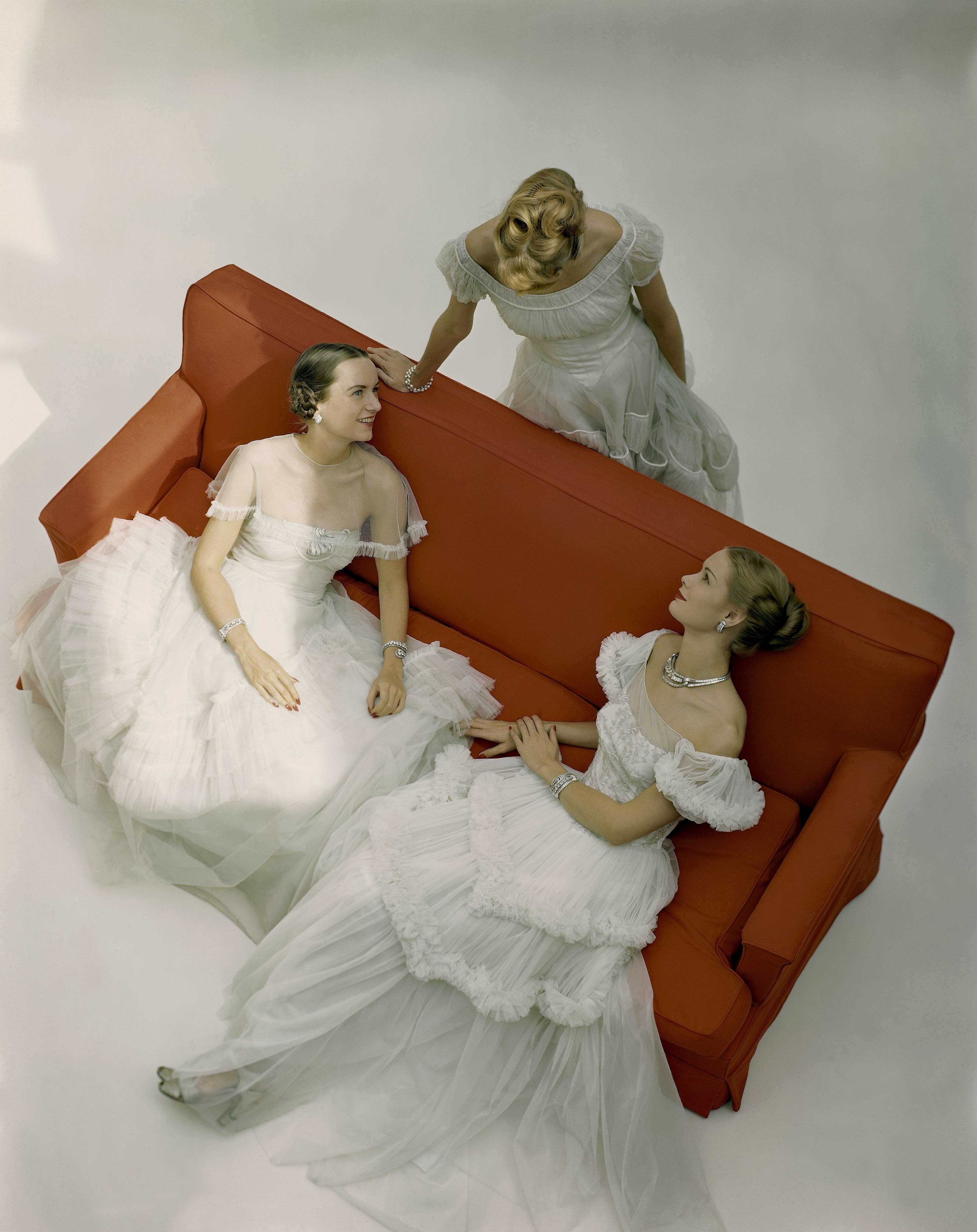Three women in elegant white gowns, one standing behind a red couch while the other two are seated, looking at each other. Names are not identifiable