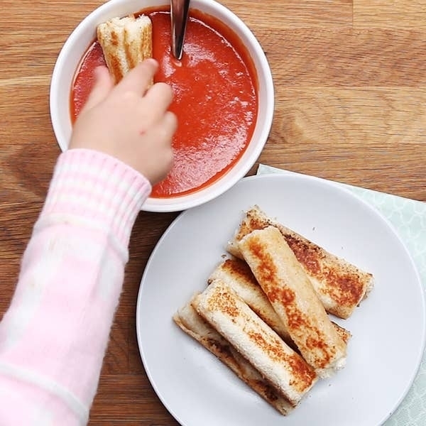 A child's hand dipping a grilled cheese stick into a bowl of tomato soup, with more grilled cheese sticks on a white plate nearby