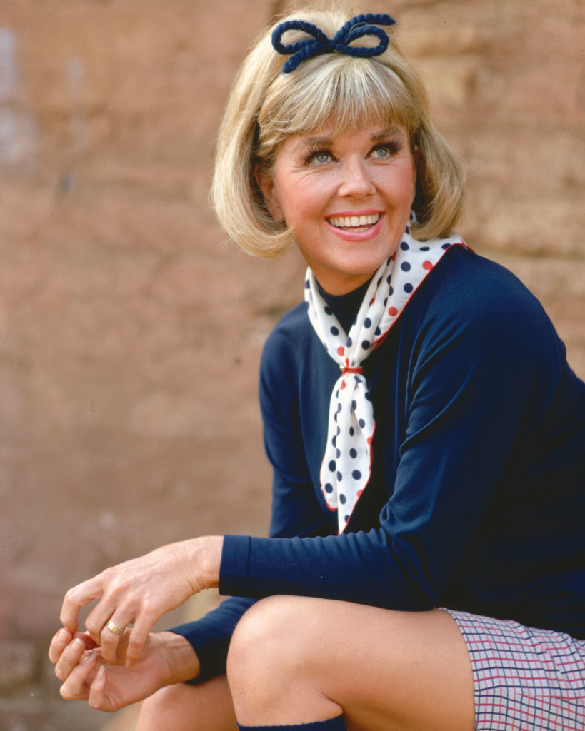 Doris Day smiling while sitting, wearing a bow hair accessory, a polka dot scarf, a long-sleeve top, and a patterned skirt